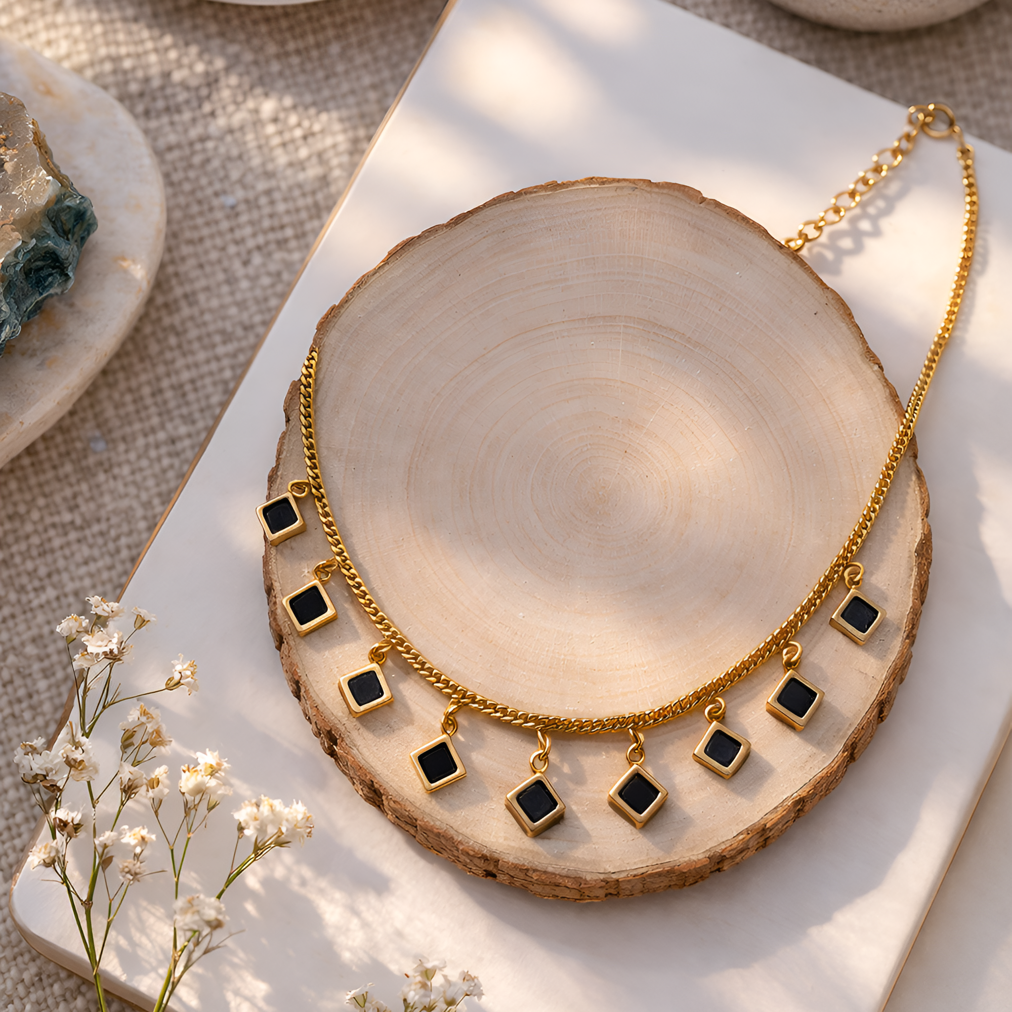Gold necklace with black square pendants on a wooden surface with flowers and stones.