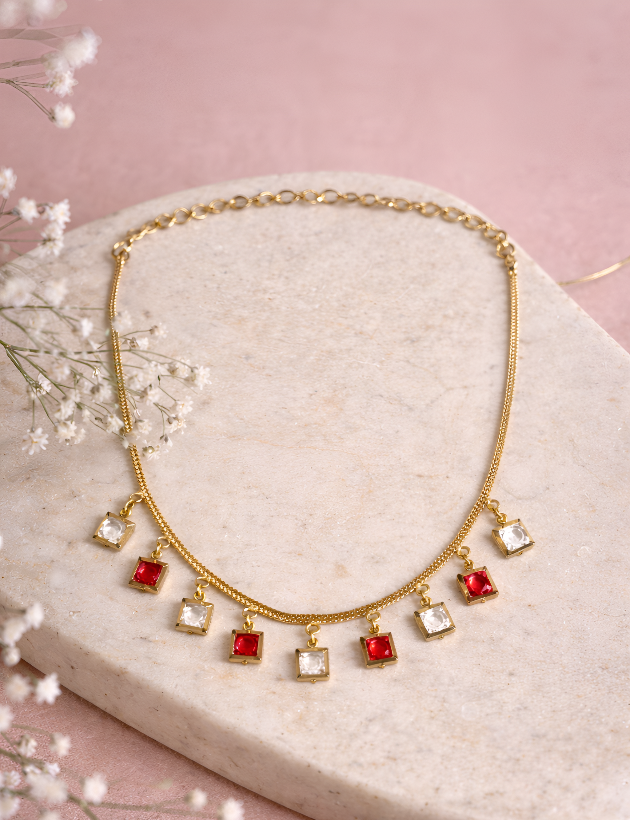 Gold necklace with red and clear square pendants on a pink background